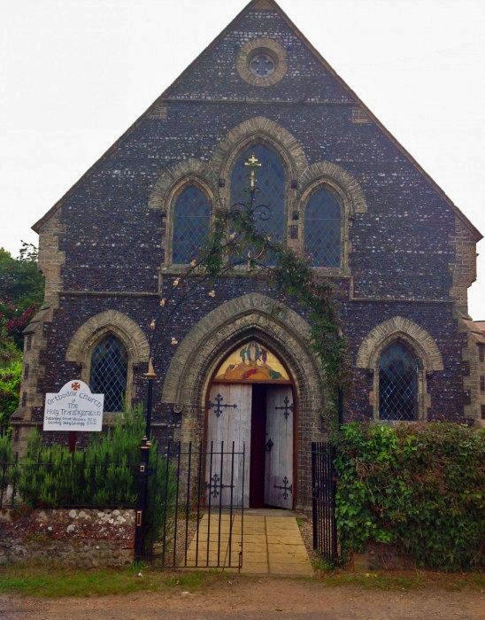 A flag stone path leads from and black iron gate to the entrance of the Orthodox Church. The building is faced with knapped flint with the arched doorway in the centre flanked by an arched window on either side and a larger window with three arches above. The glass is diamond-paned. The double wooden doors have decorative black hinges and is surmounted by a stylised depiction of the transfiguration of Christ with a gold background.