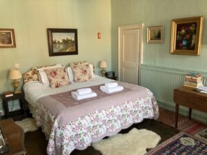 View of a double bed with lavender counterpane, two bundles of white towels. On the walls are framed paintings including a river landscape and a floral still life.