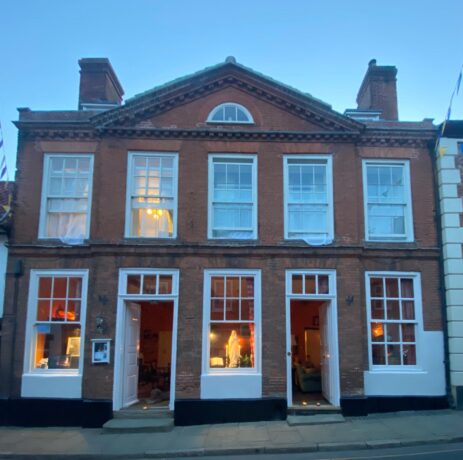 Frontage of the Old Bakehouse lit up at dusk. The building is a two storey redbrick house a chimney stack on both sides of a triangular pediment. There are five large sash windows on the upper level and three on the ground floor with two doors with steps down to the flagstone pavement. The house presents a welcoming prospect with warm light shining through the windows and open doors.