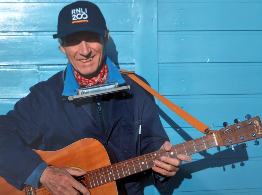 Bob, dressed in navy blue with a baseball cap shading his eyes from the sun, is strumming his acoustic guitar in front of a turquoise blue background